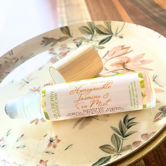 A roll-on perfume oil bottle labeled 'Honeysuckle Jasmine & Sea Mist' by Soulberry Apothecary, resting on a floral-patterned ceramic plate with its bamboo cap beside it.