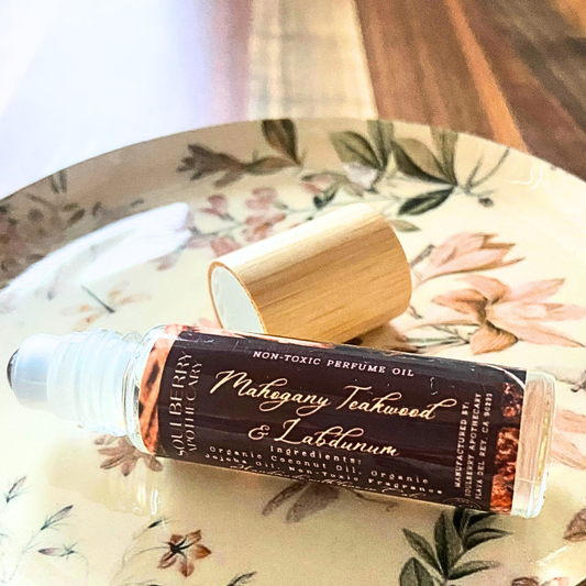 A clear glass rollerball bottle of perfume oil with a bamboo cap resting on a floral ceramic plate. The label reads 'Mahagany Teakwood & Labdanum' and 'Soulberry Apothecary'